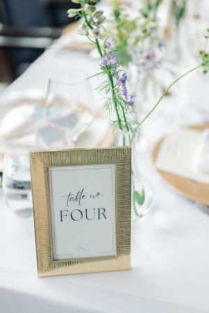 Gold-framed table number cards with delicate purple flowers at a wedding reception.