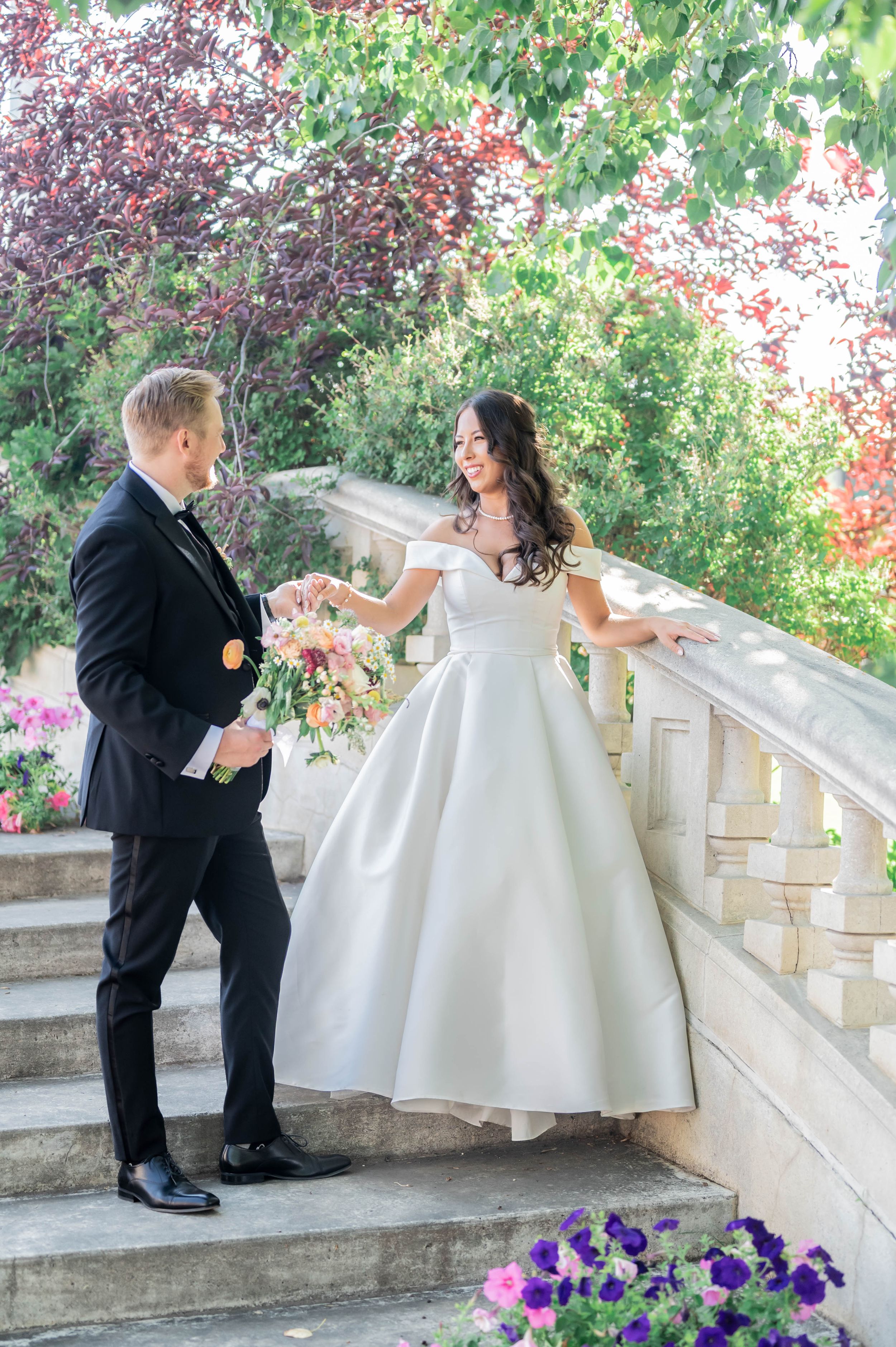 Couple in formal wedding attire playfully posing on stone steps with purple flowers below.