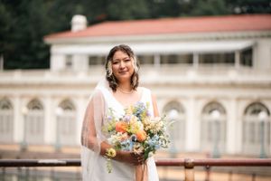 A bride holding a colorful bouquet poses against classic architecture with a red roof.
