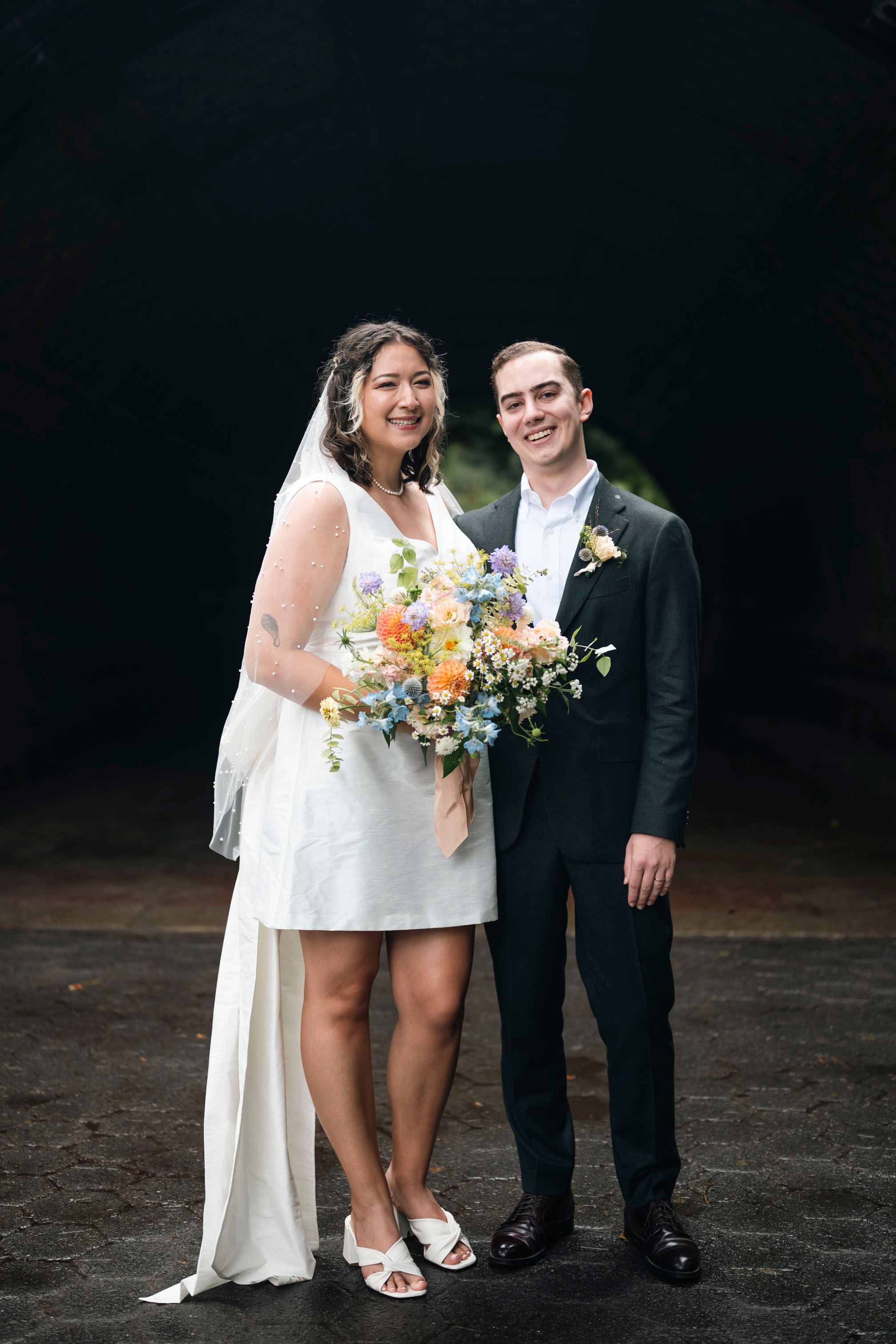 A full-length wedding portrait featuring a short dress and traditional black tuxedo.