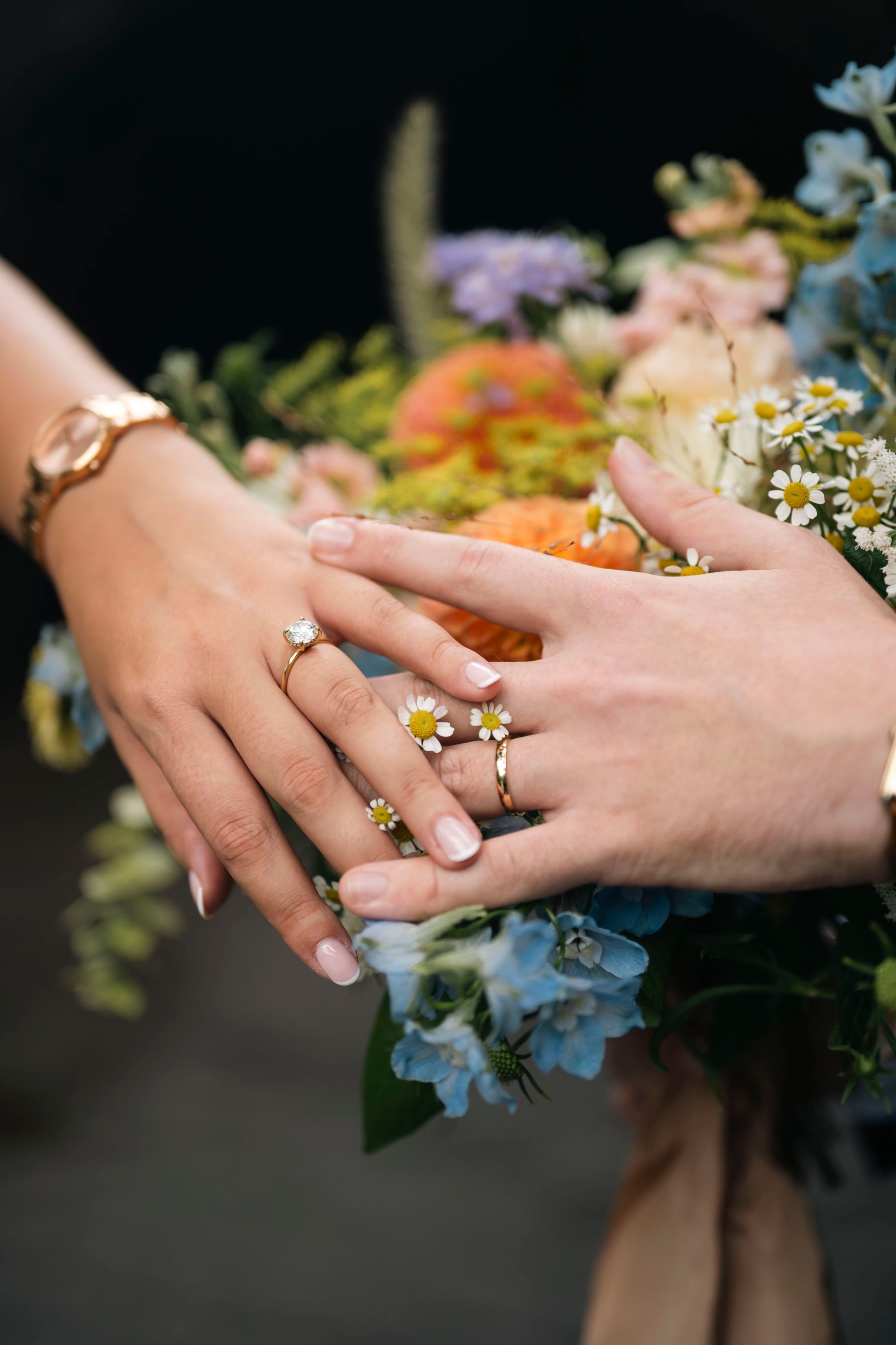 Close-up of hands showing wedding rings against a backdrop of blue and peach flowers in a bridal bouquet.