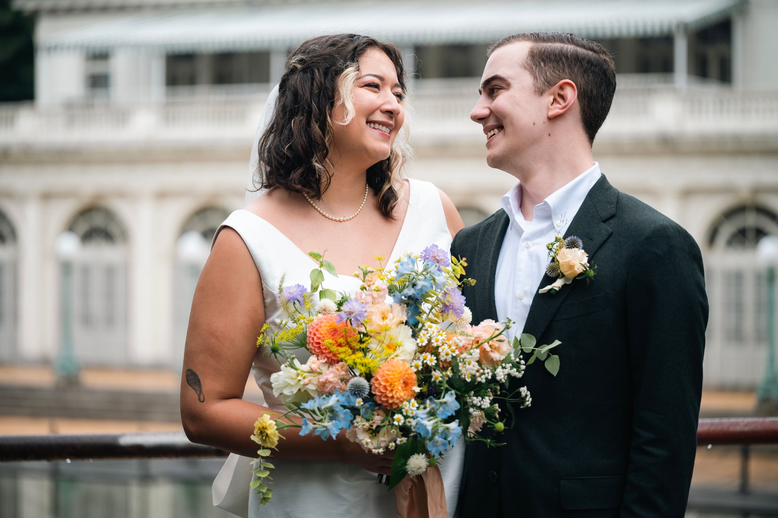 A joyful wedding couple sharing a laugh while holding a vibrant bouquet of orange and white flowers against a classic building.