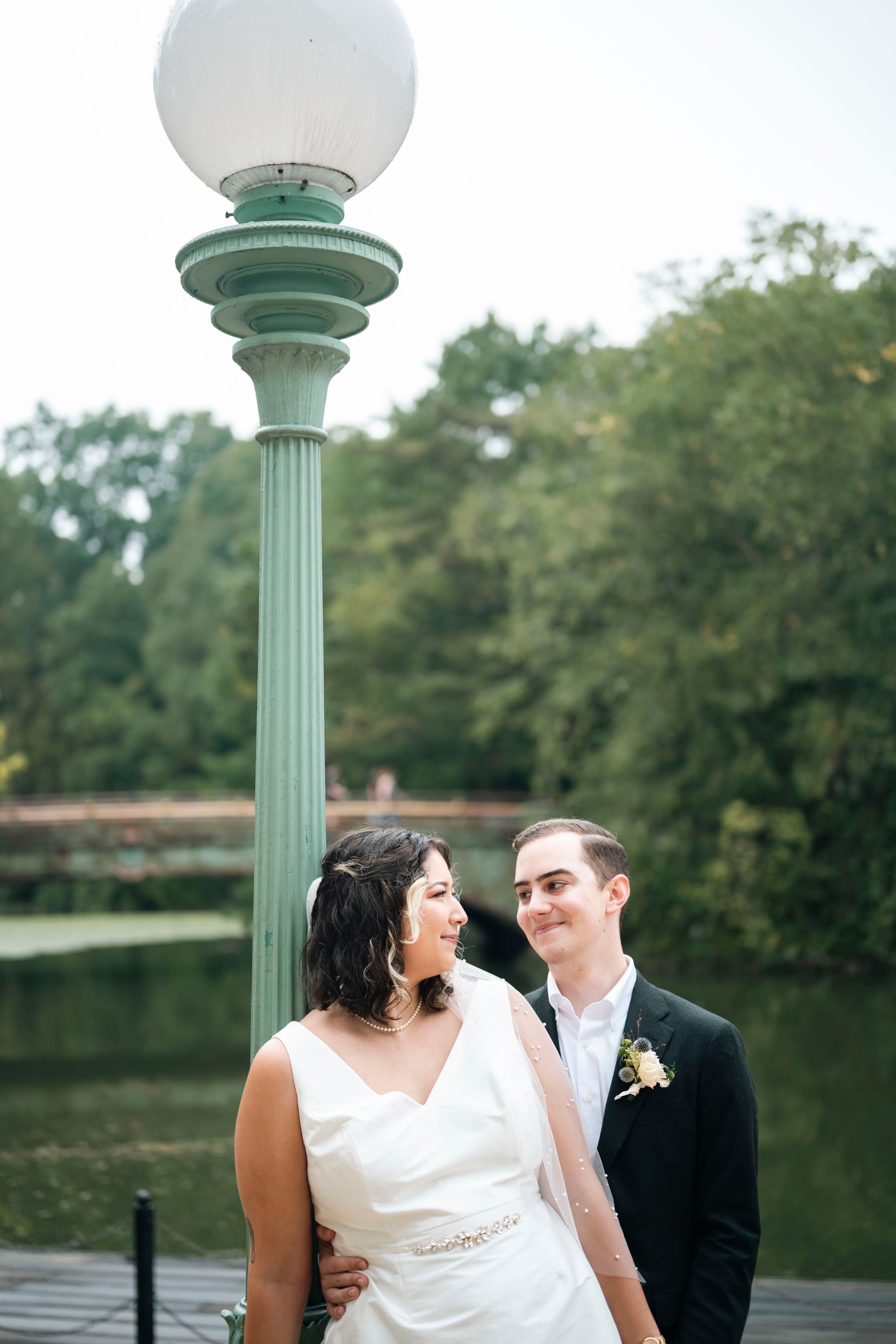 A wedding couple poses beneath a vintage green lamp post near a serene pond.