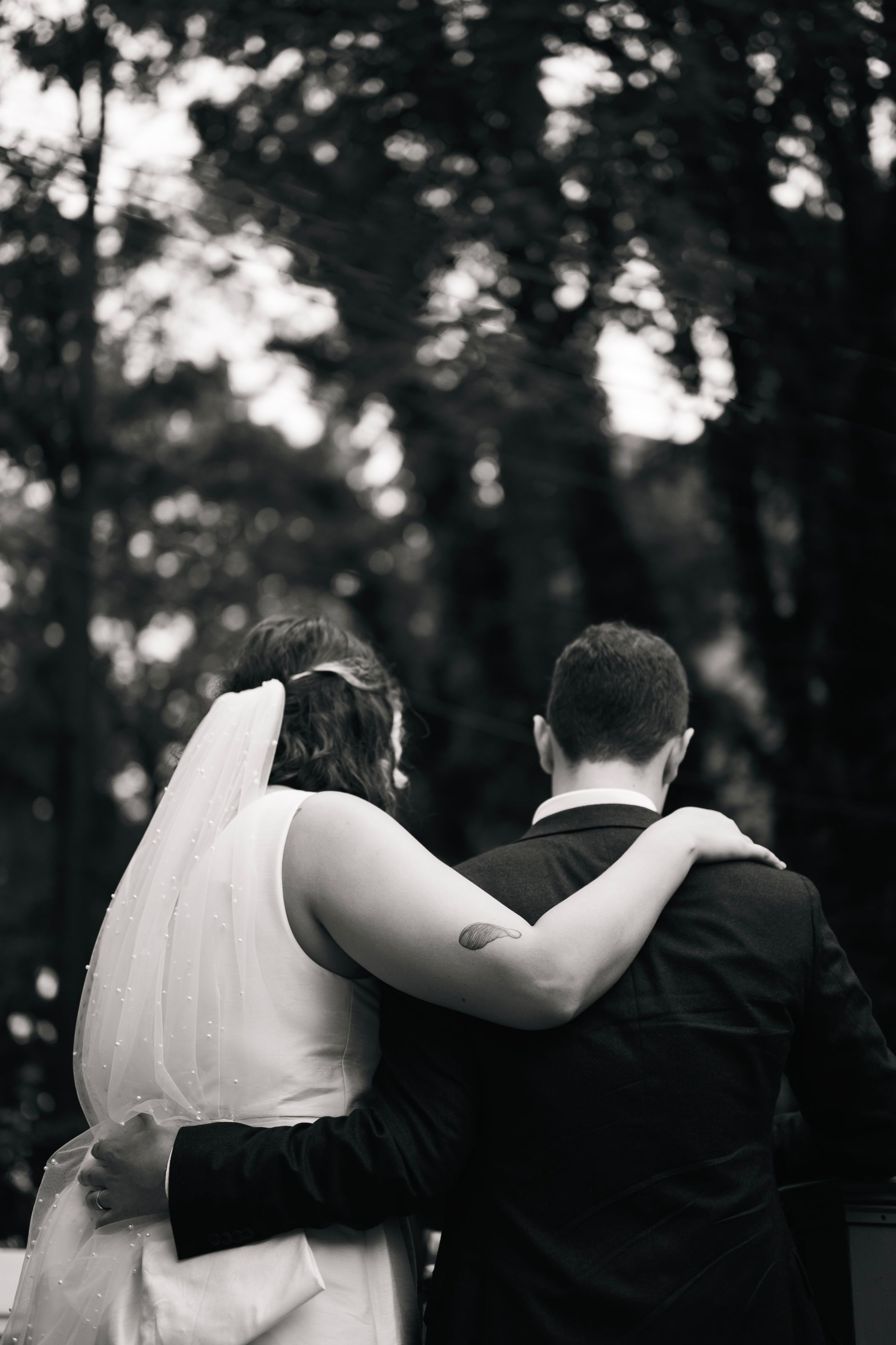Black and white photo of a couple embracing during their wedding ceremony in a wooded setting.