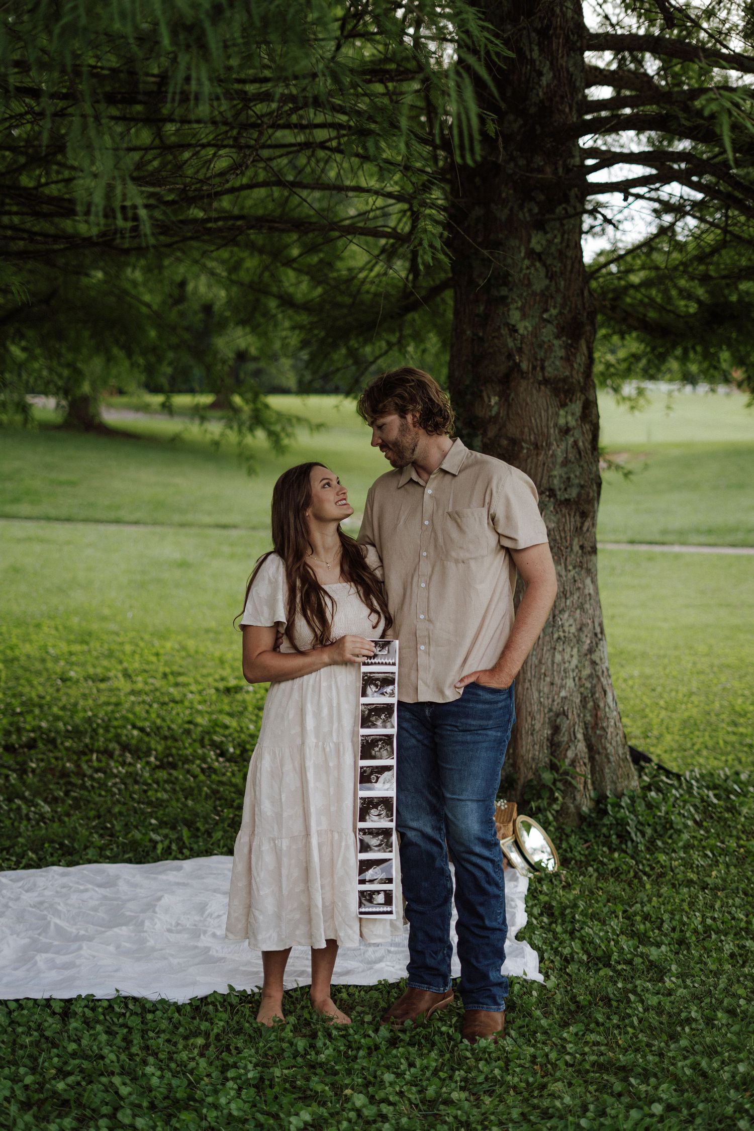 Couple standing under a tree holding ultrasound photos, announcing pregnancy outdoors on a green lawn.
