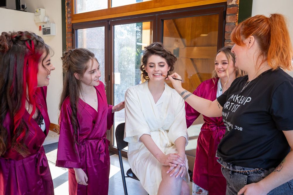 Bride in white robe gets makeup done by artist while bridesmaids in purple robes smile during wedding prep.