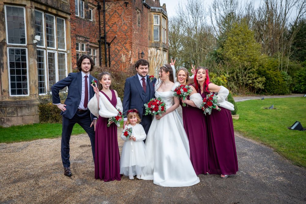 Wedding party poses outside brick manor, bride in white gown with groom, bridesmaids in burgundy dresses holding red bouquets.