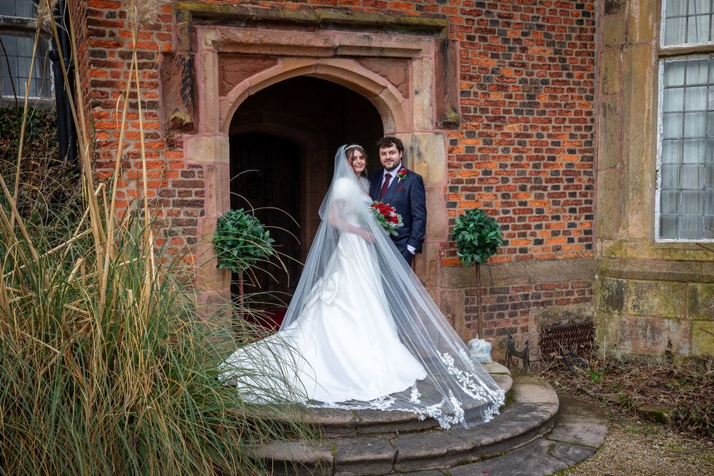 Bride in flowing white gown with long veil and groom in suit pose on stone steps outside a red brick manor.
