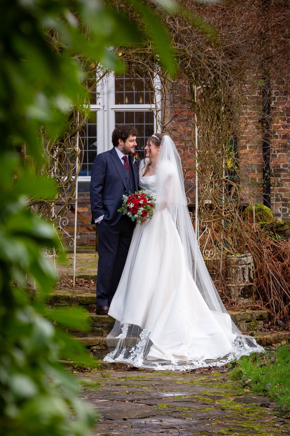 Bride and groom stand together in a garden, bride wearing white gown with veil holding red bouquet.