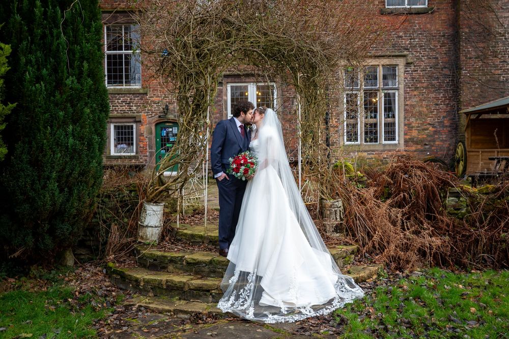 Bride and groom kissing on stone steps outside a red brick manor house, bride in white gown with lace veil.