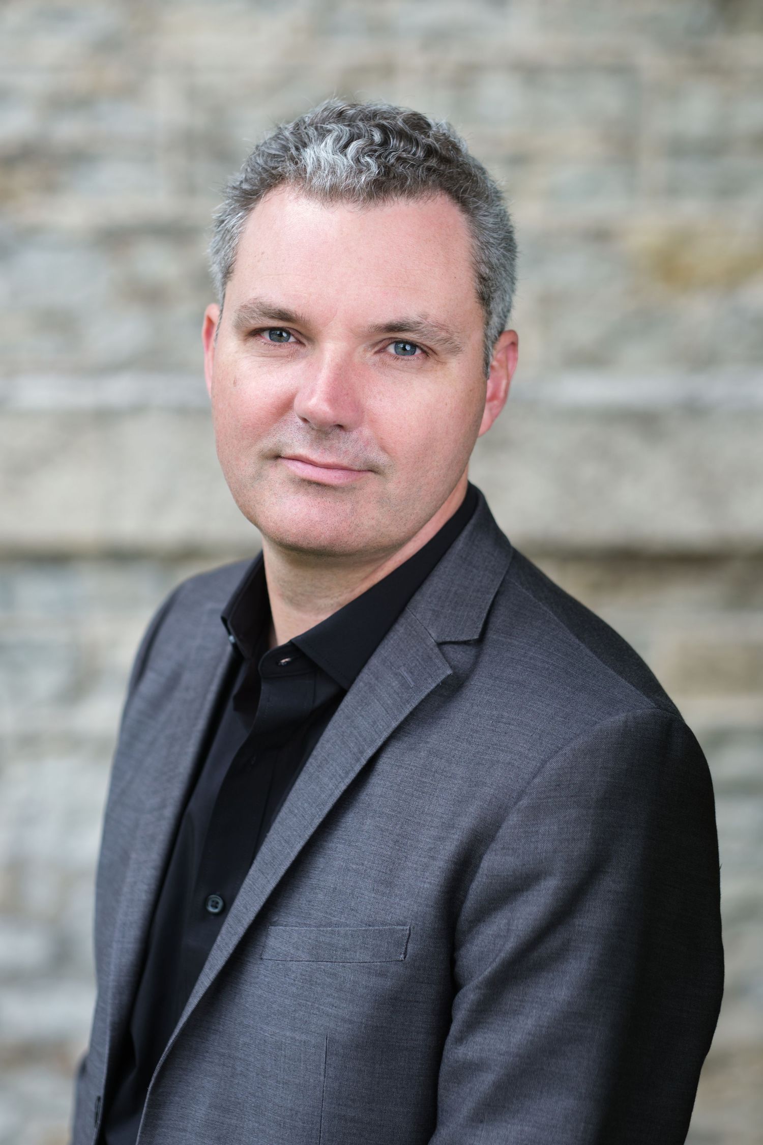 A musician stands for a headshot in a suit in front of a textured brick wall in Appleton, Wisconsin