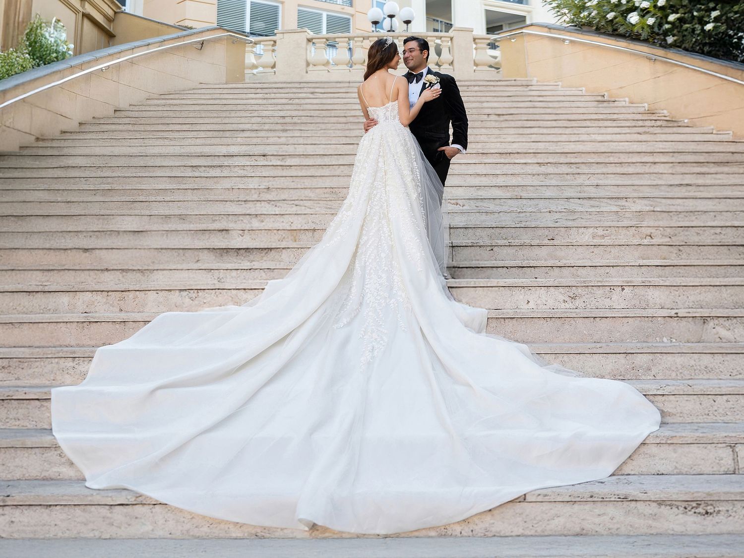 Bride and groom standing on grand outdoor staircase, bride wearing elegant white gown with long flowing train.