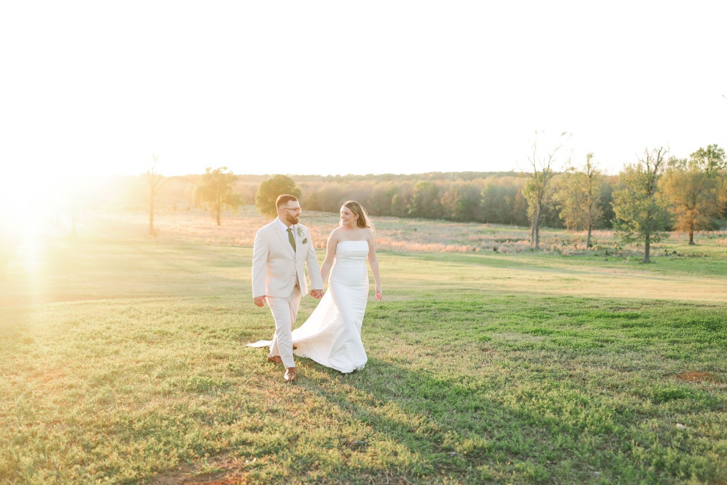 Bride and groom walking hand in hand through a sunlit green field during golden hour at an outdoor wedding.