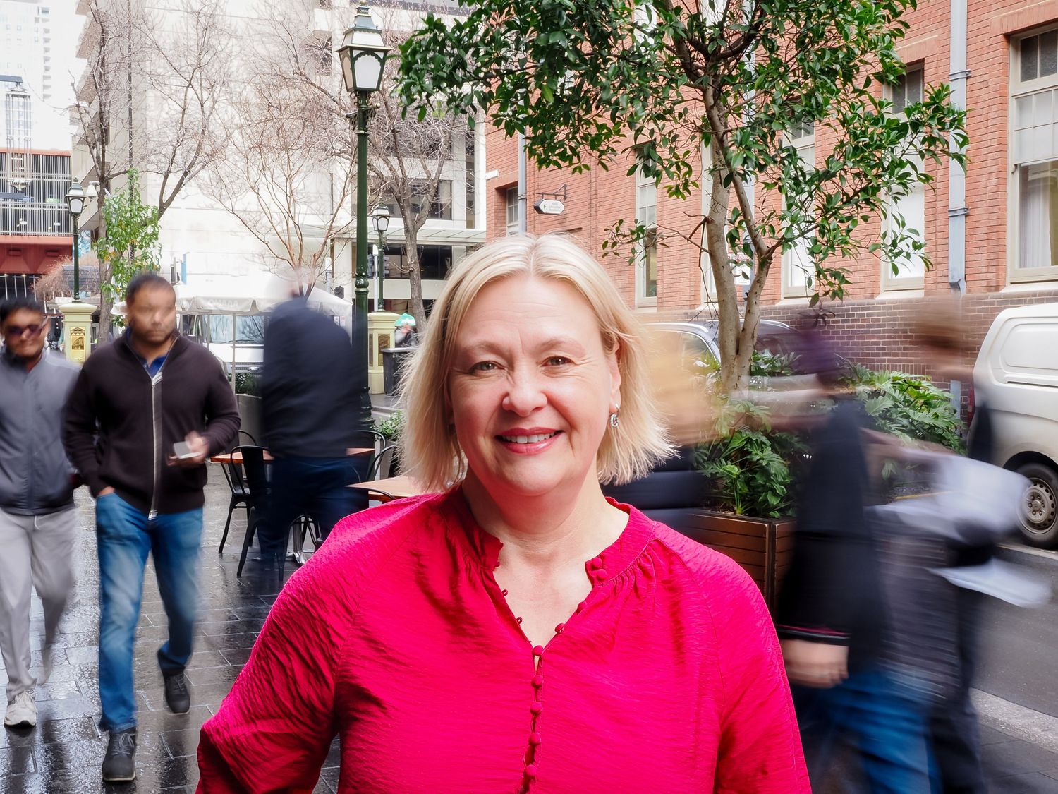 Smiling blonde woman in red top on busy urban street with blurred pedestrians and green trees in background.