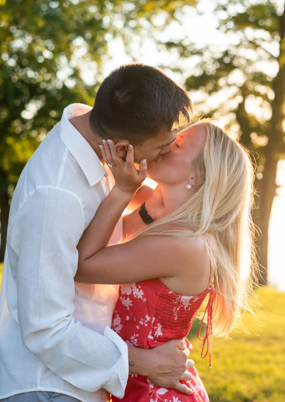 Couple sharing a romantic kiss outdoors at golden hour, woman in red floral dress embracing man in white shirt.