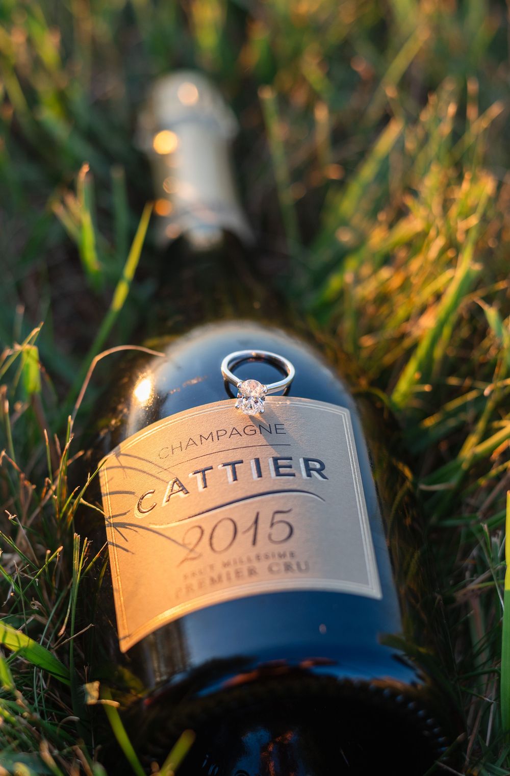 Diamond engagement ring resting on a Cattier 2015 Premier Cru Champagne bottle lying in green grass.