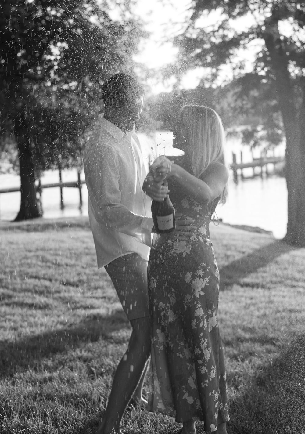 Black and white photo of a couple popping champagne outdoors near a waterfront, surrounded by trees.