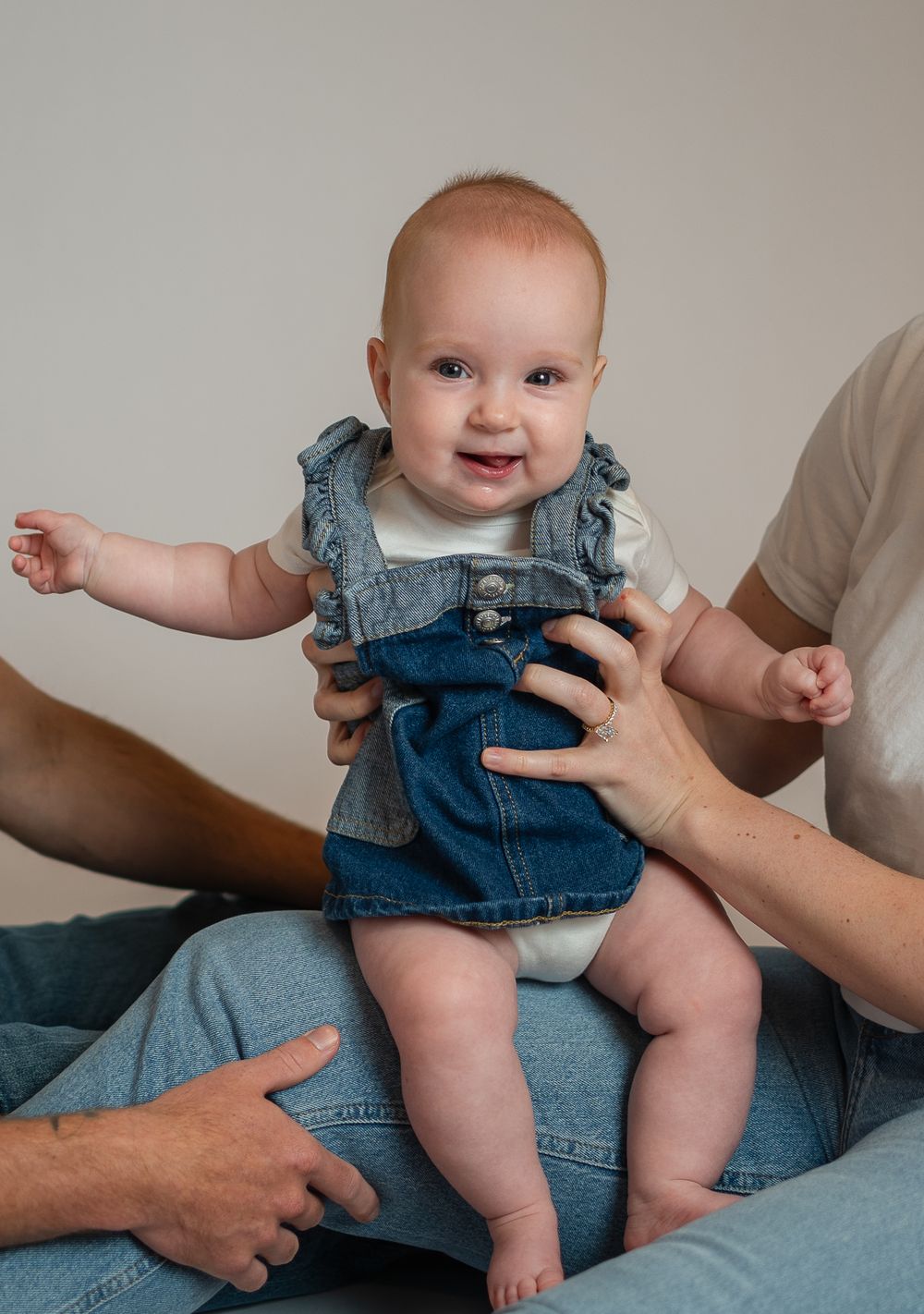 Smiling baby in denim overalls being held up by parents hands, wearing a white onesie underneath.