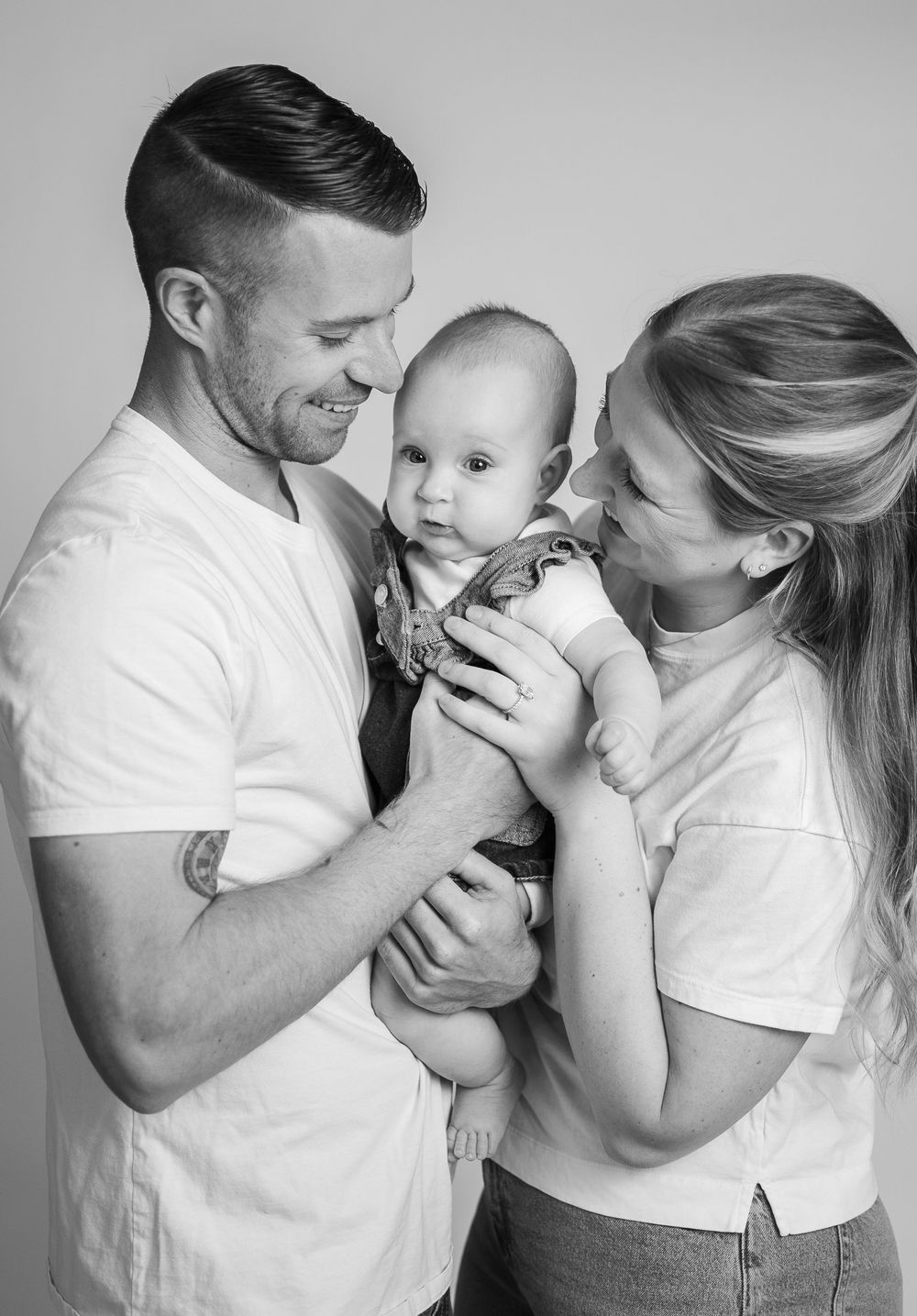 Black and white photo of smiling parents holding their newborn baby against a light studio background.
