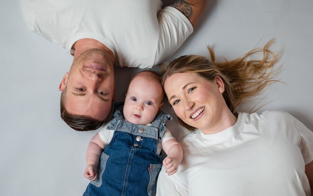 Overhead view of smiling parents lying down with their baby dressed in denim overalls on a white background.