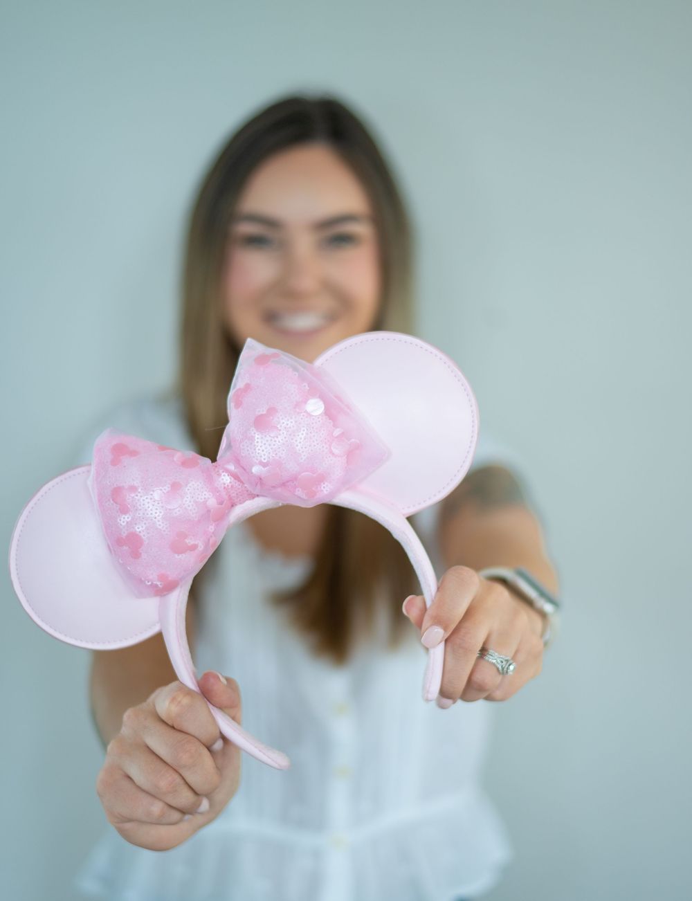 Woman smiling and holding pink Minnie Mouse ears headband with sequin bow toward camera.