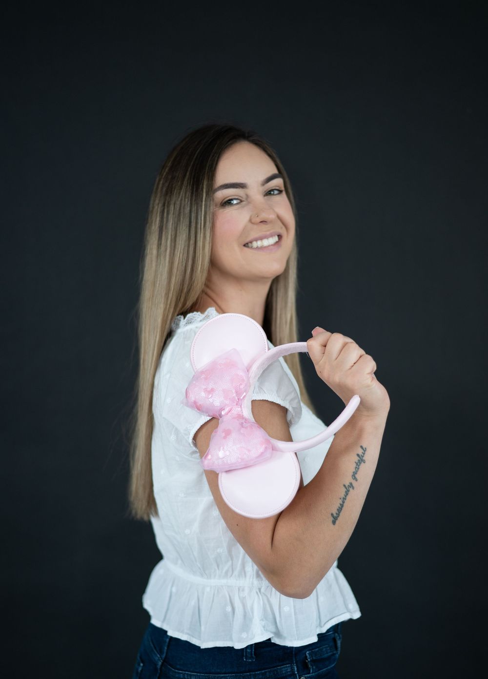 Smiling woman with ombre hair holding a pink Minnie Mouse ear headband, wearing a white top with a script tattoo on her arm.