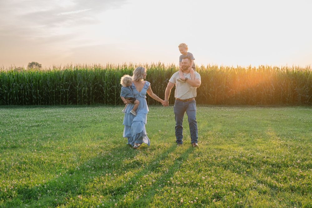 Family walking in a green field at sunset near a cornfield, parents holding children in warm golden light.