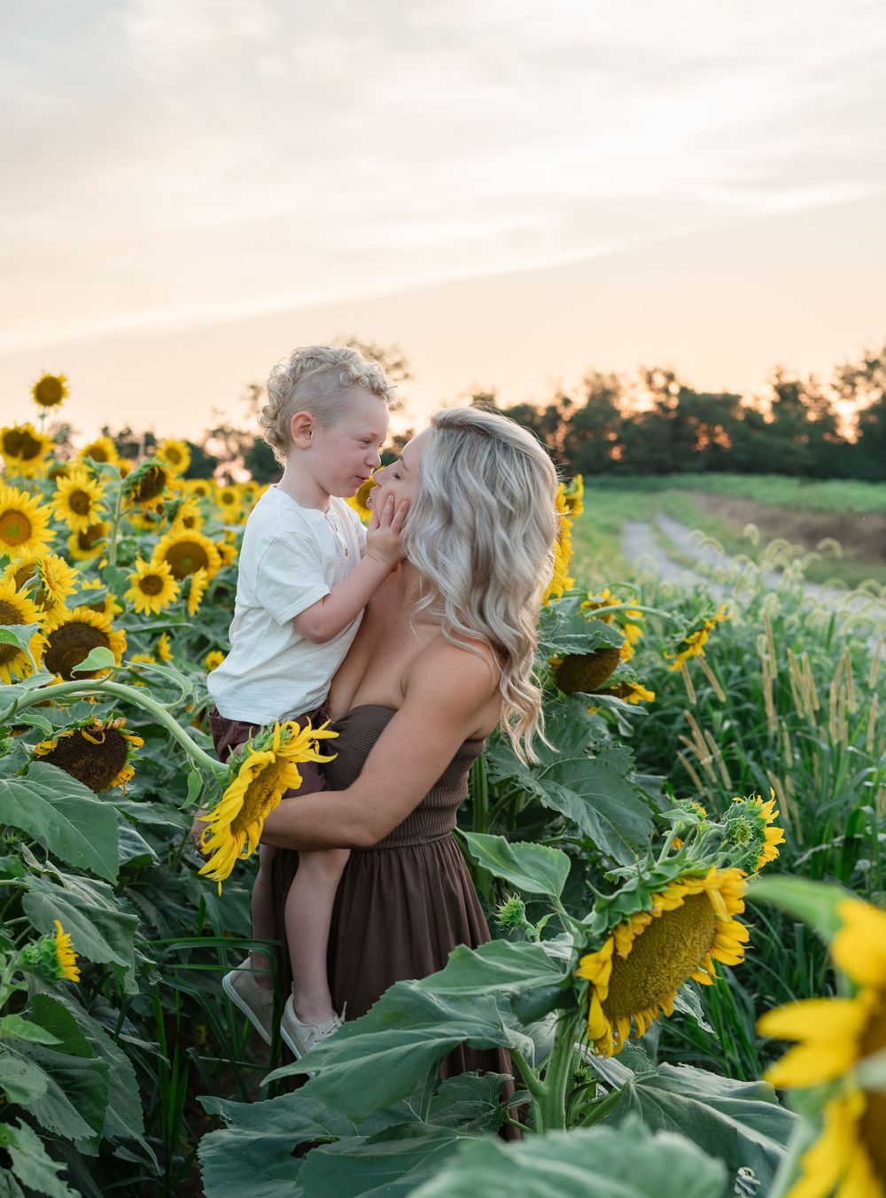 A mother holds her toddler in a sunflower field at golden hour, both sharing a tender moment at sunset.