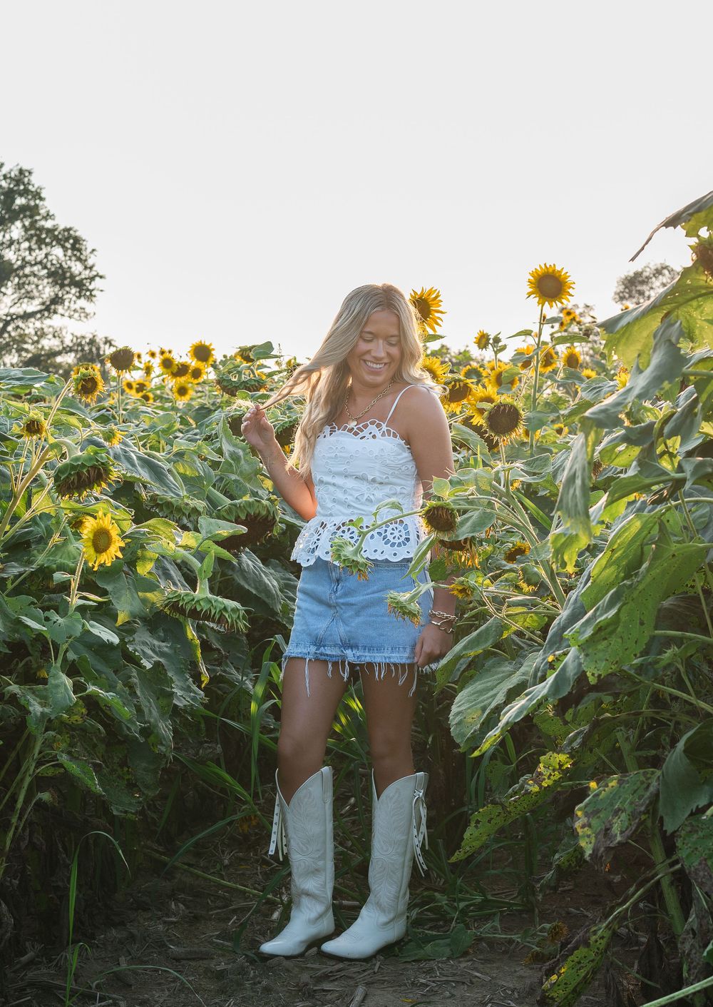 Woman in white lace top, denim fringe skirt, and white cowboy boots posing in a vibrant sunflower field.