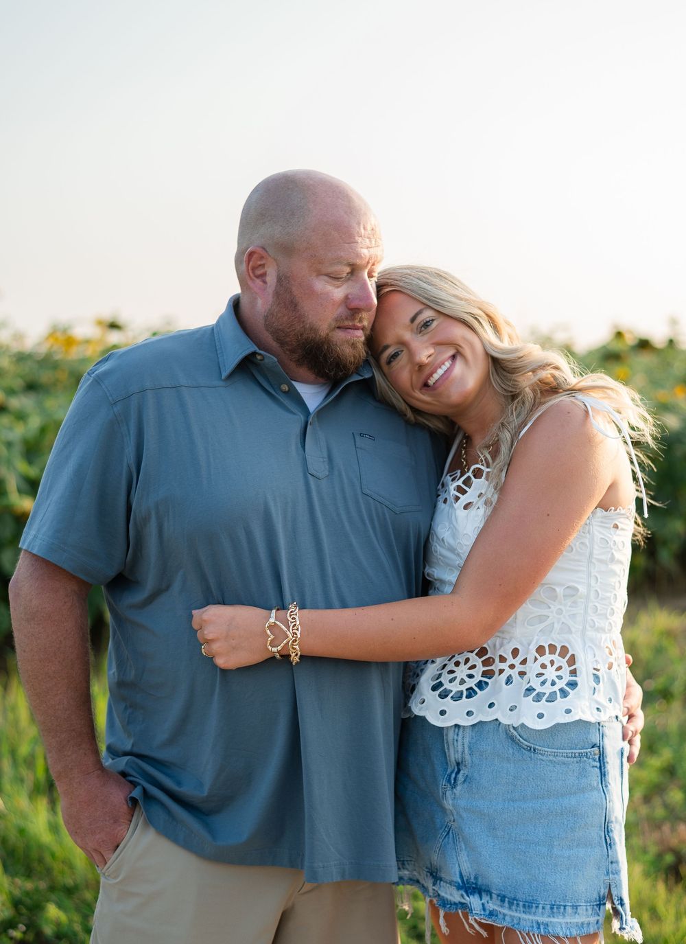 A couple poses romantically in a sunflower field, woman in lace top and denim skirt leaning on man in blue polo.