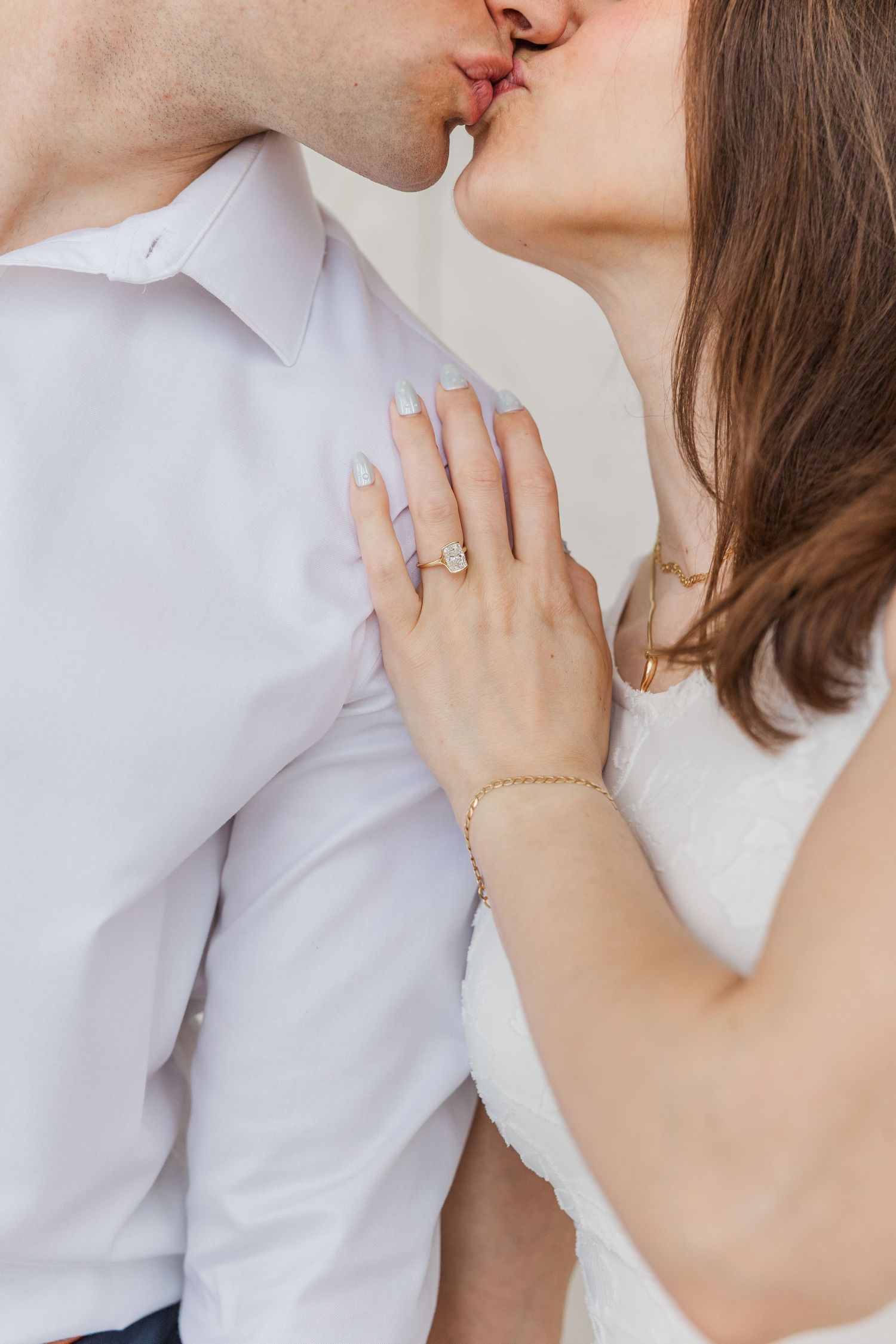 Couple sharing a romantic kiss, woman displaying diamond engagement ring and gold bracelet on mans chest.