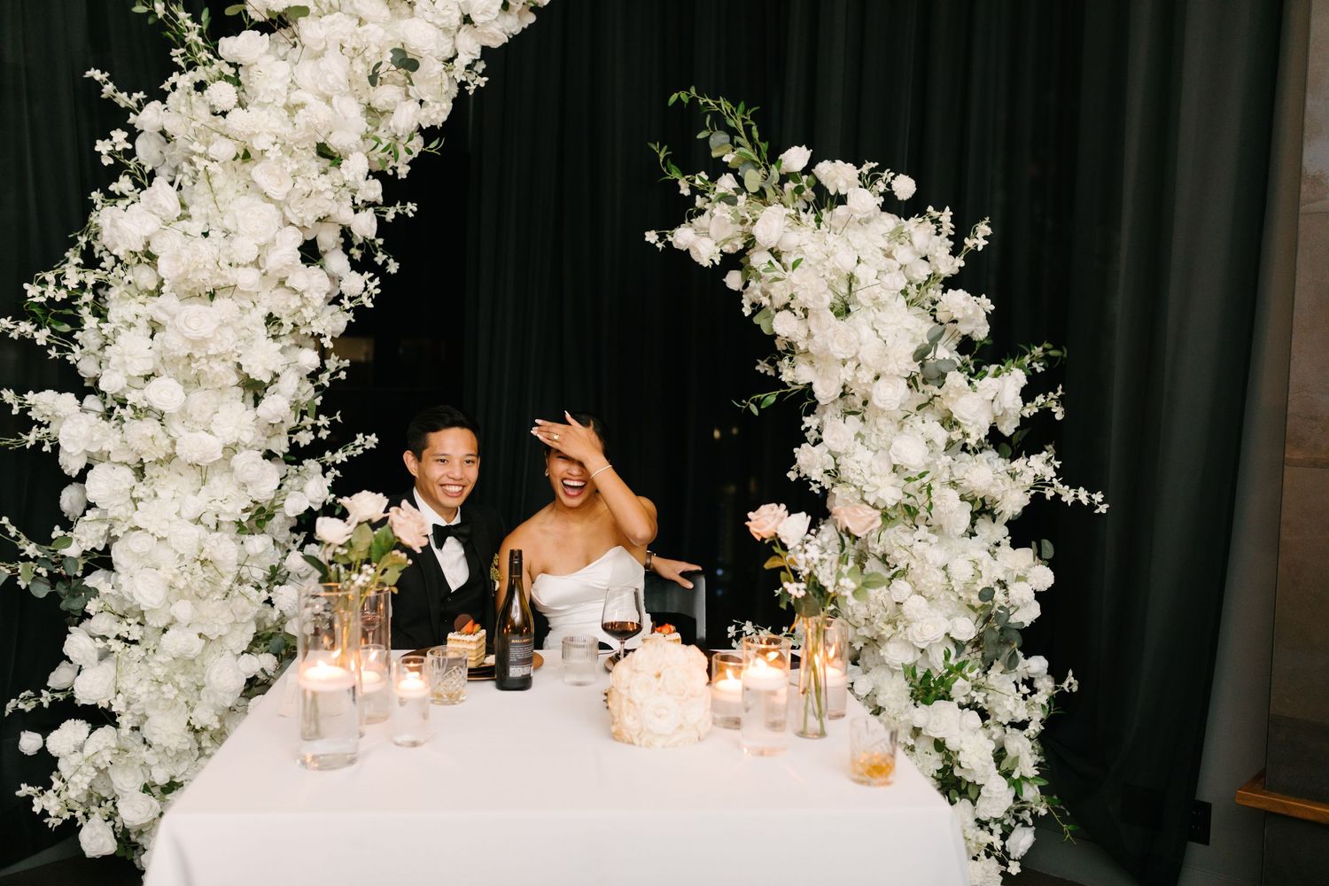 Newlyweds seated at a head table beneath a tall white floral arch.