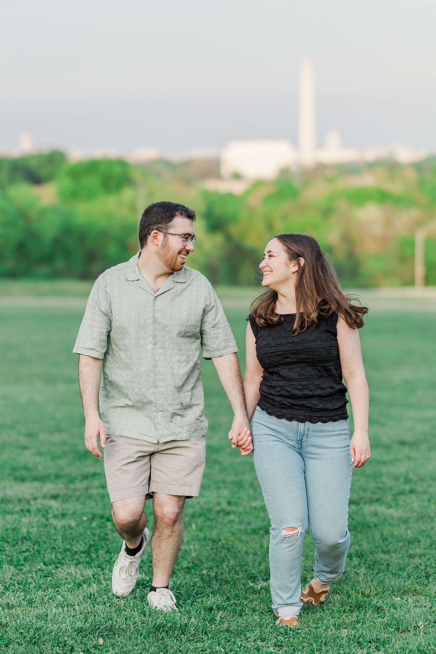 Couple holding hands and smiling at each other while walking on green grass with Washington Monument in background.