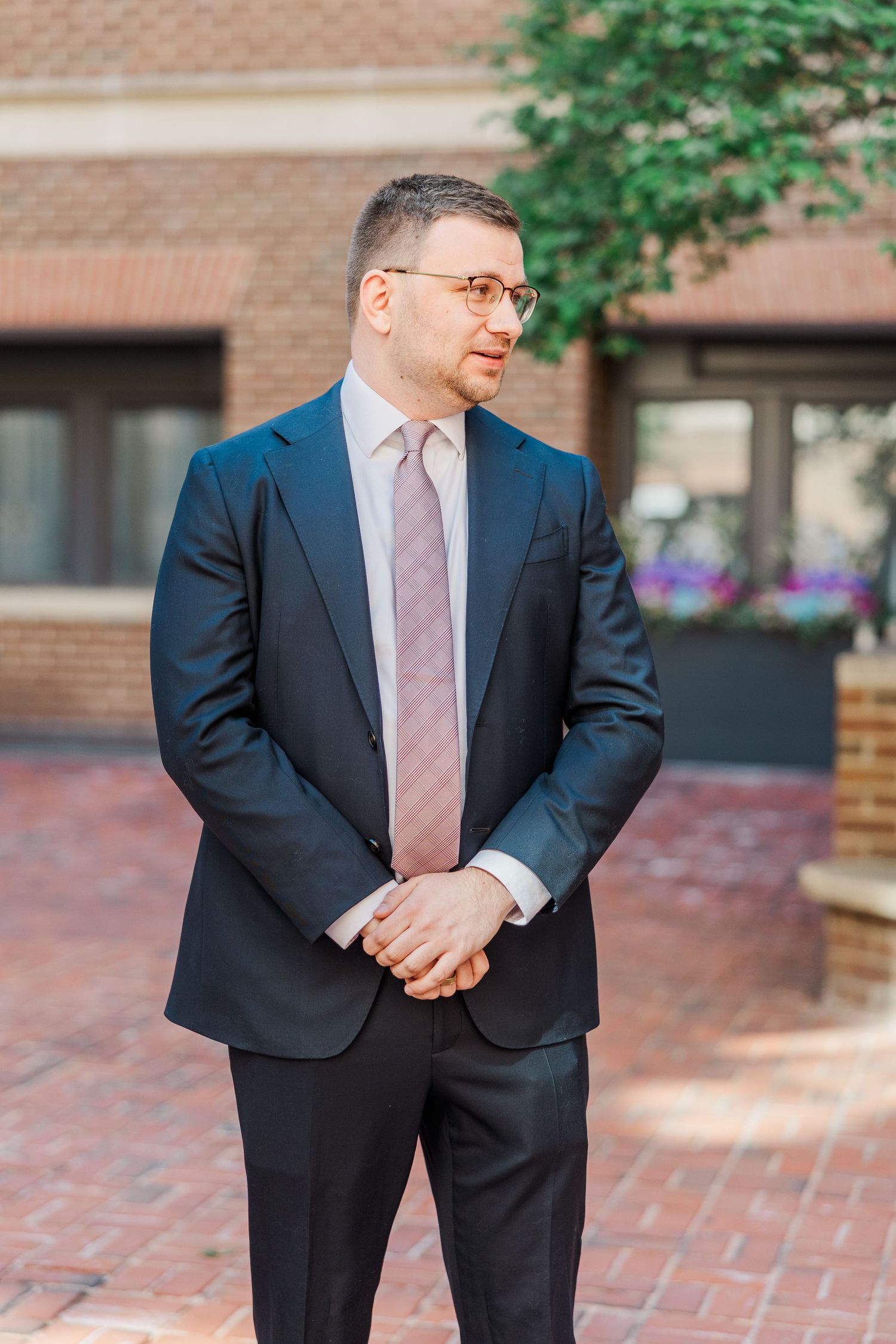 A well-dressed man in a dark navy suit and pink tie stands outdoors on a brick patio, wearing glasses.
