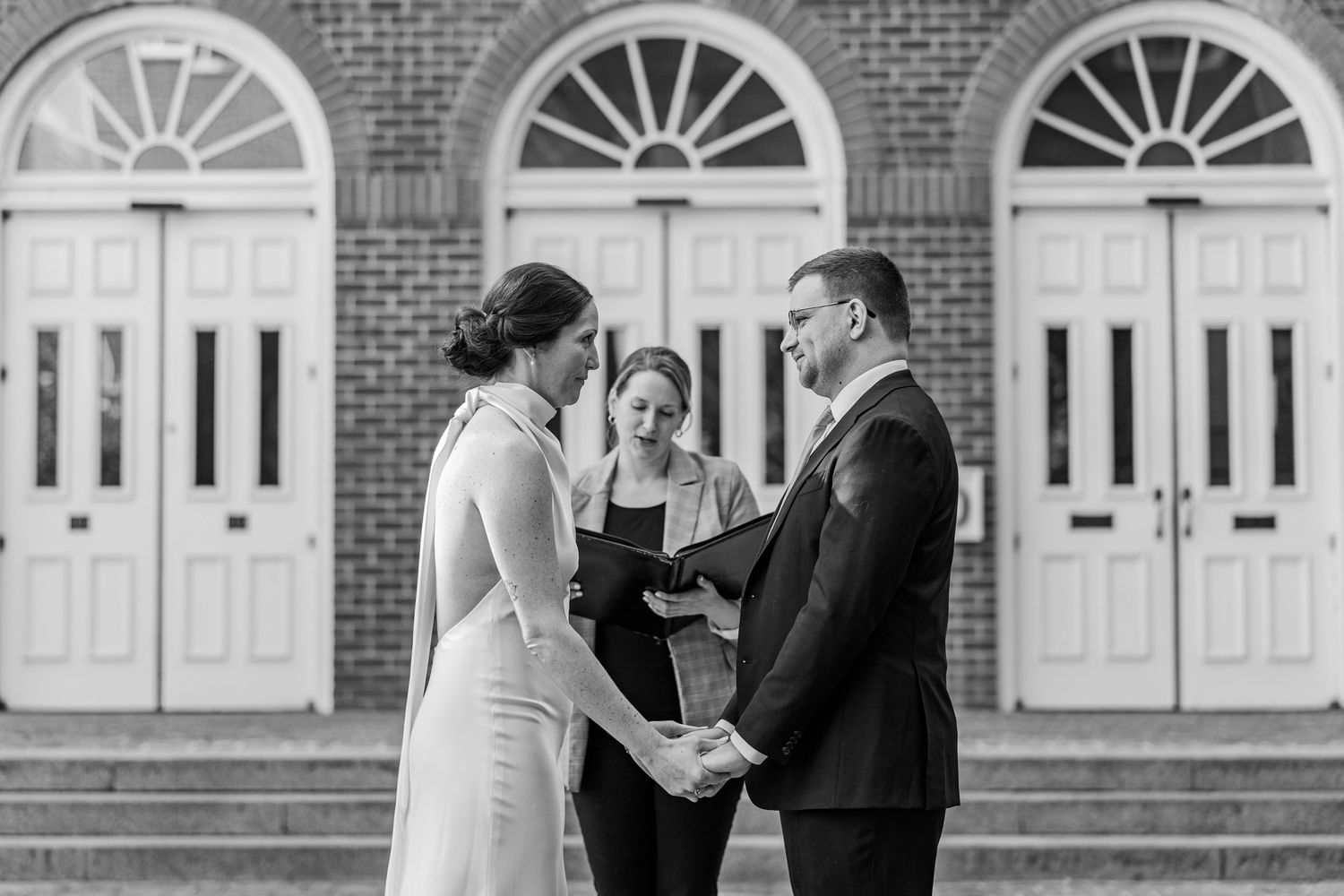 Black and white wedding ceremony photo of couple exchanging vows outdoors before brick building with arched windows.
