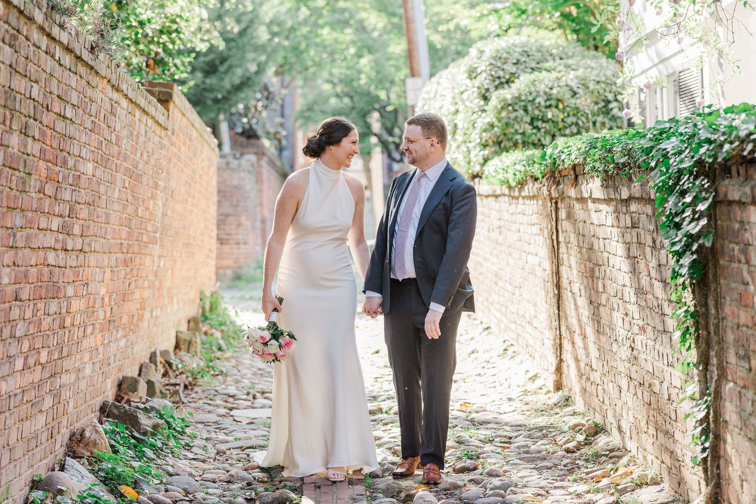 Bride and groom share a romantic kiss in a brick alleyway, bride holding a pink bouquet in a white gown.