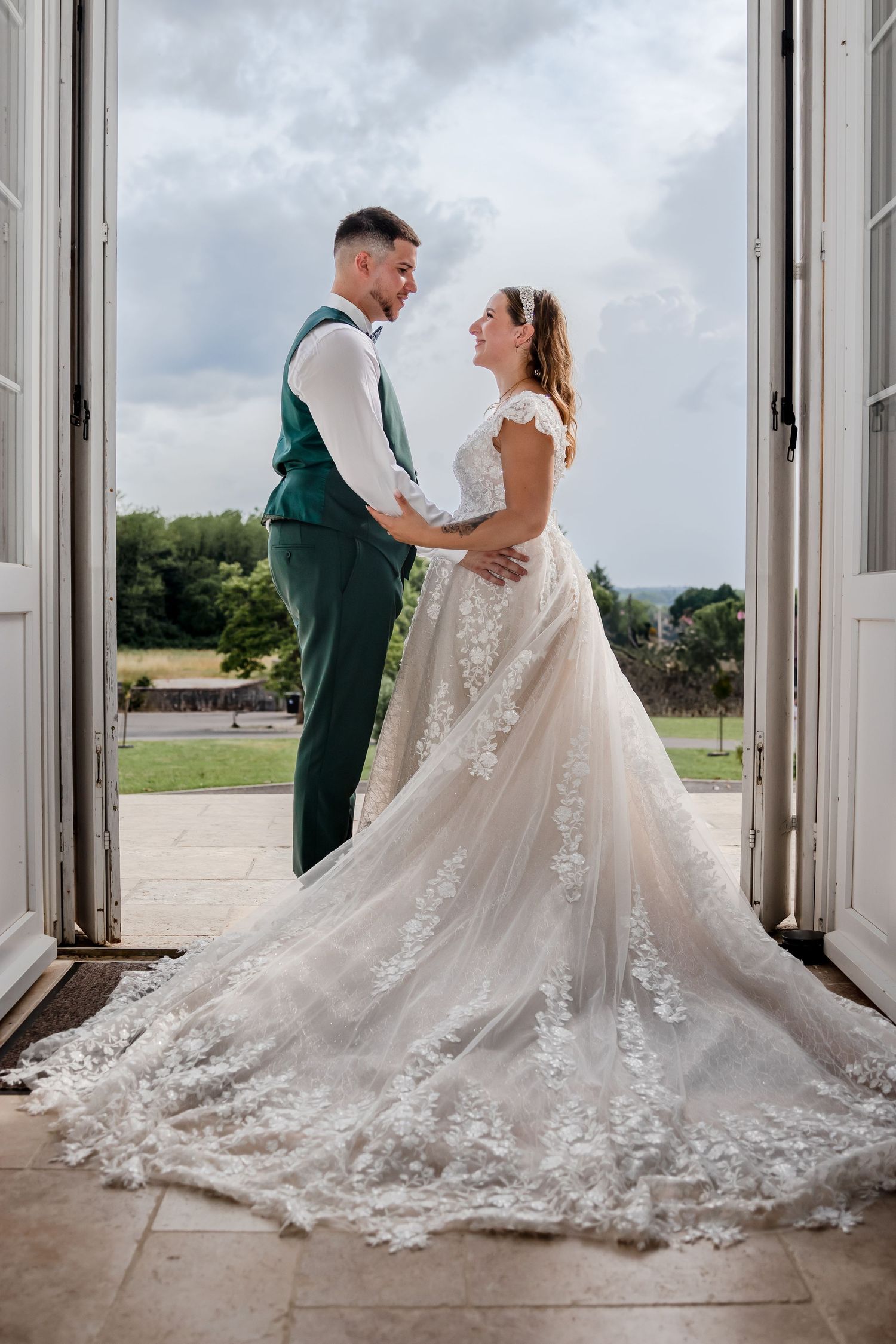 Bride in lace gown and groom in green suit embrace in open doorway with scenic garden views on their wedding day.