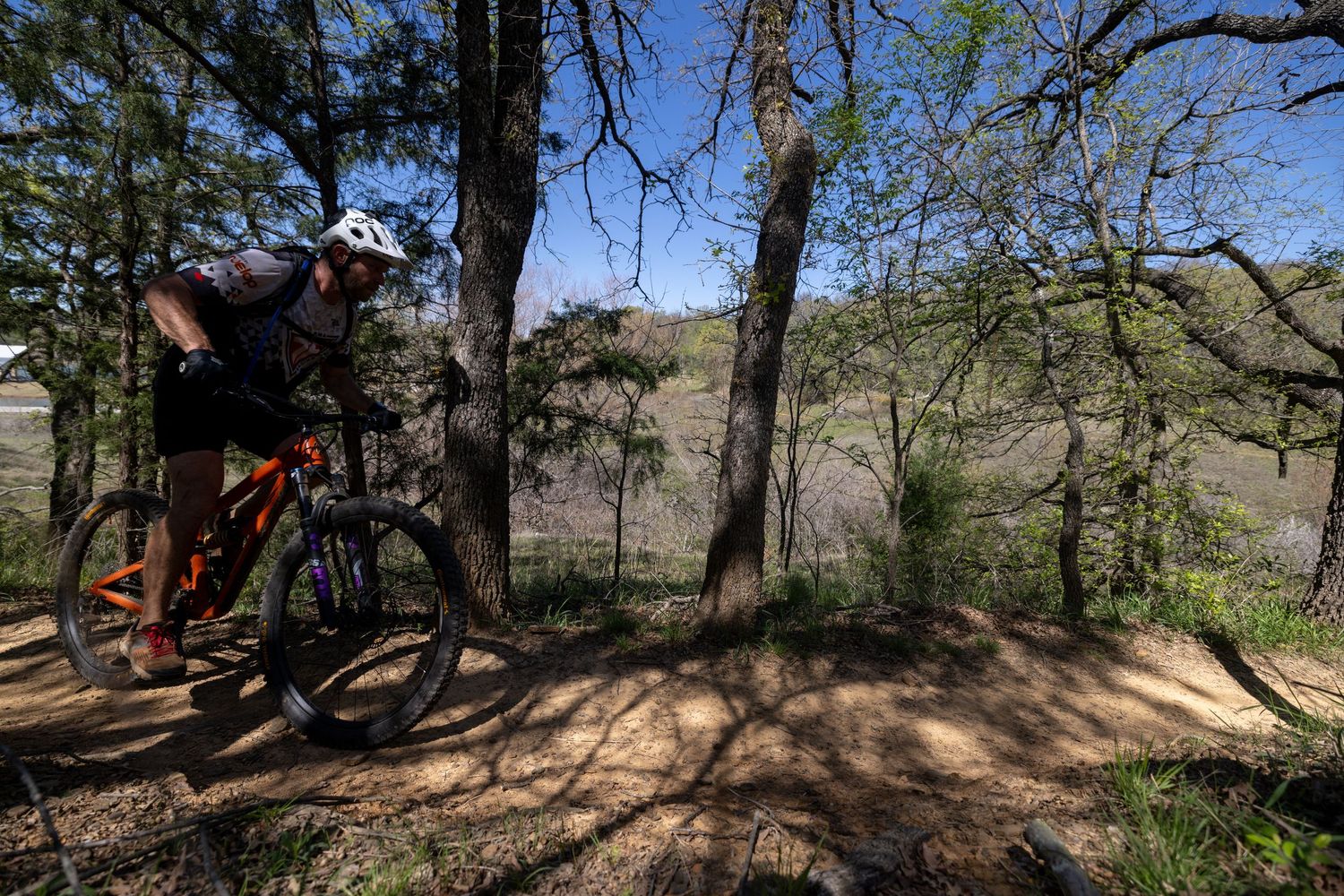 A winding nature trail with a wooden footbridge meanders through a lush Texas Hill Country landscape under a clear blue sky.