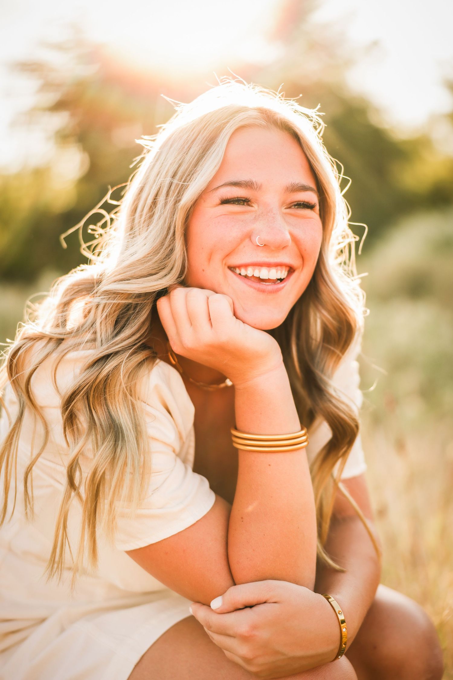 Smiling blonde teenage girl with wavy hair wearing a white dress and gold bracelets, sitting outdoors in golden sunlight.