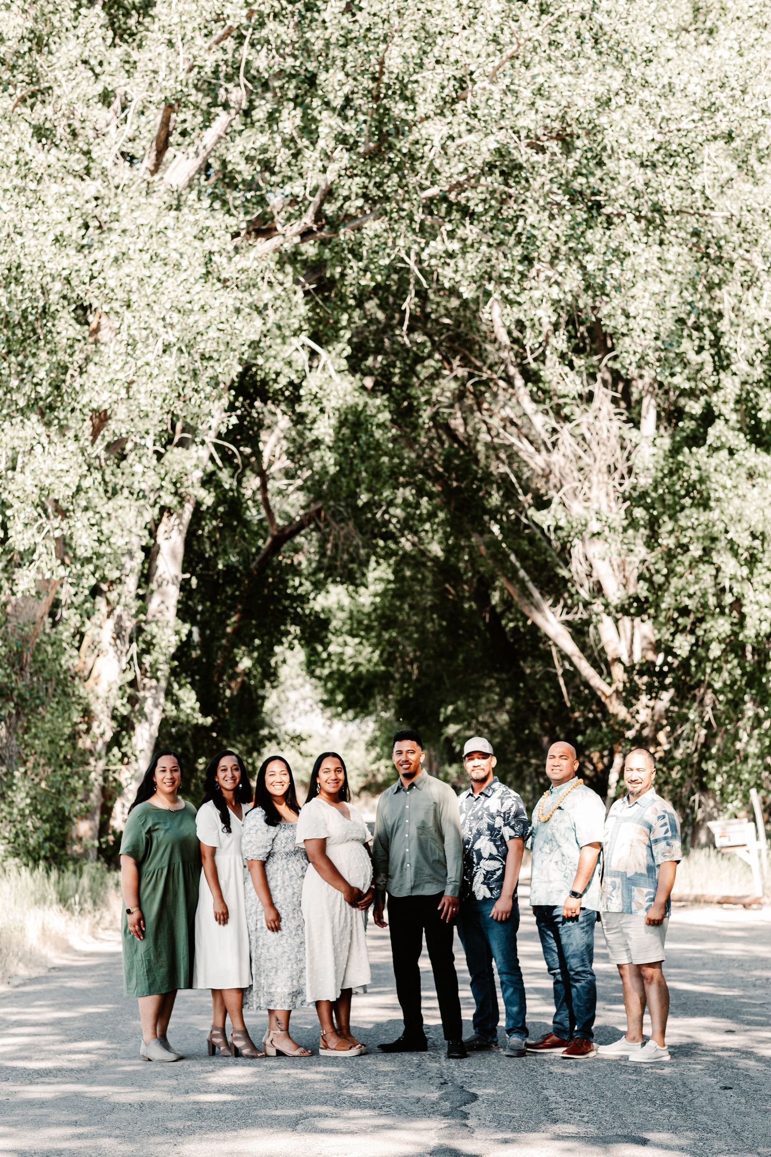 A group of eight people posing together on a sunlit path surrounded by lush green trees.