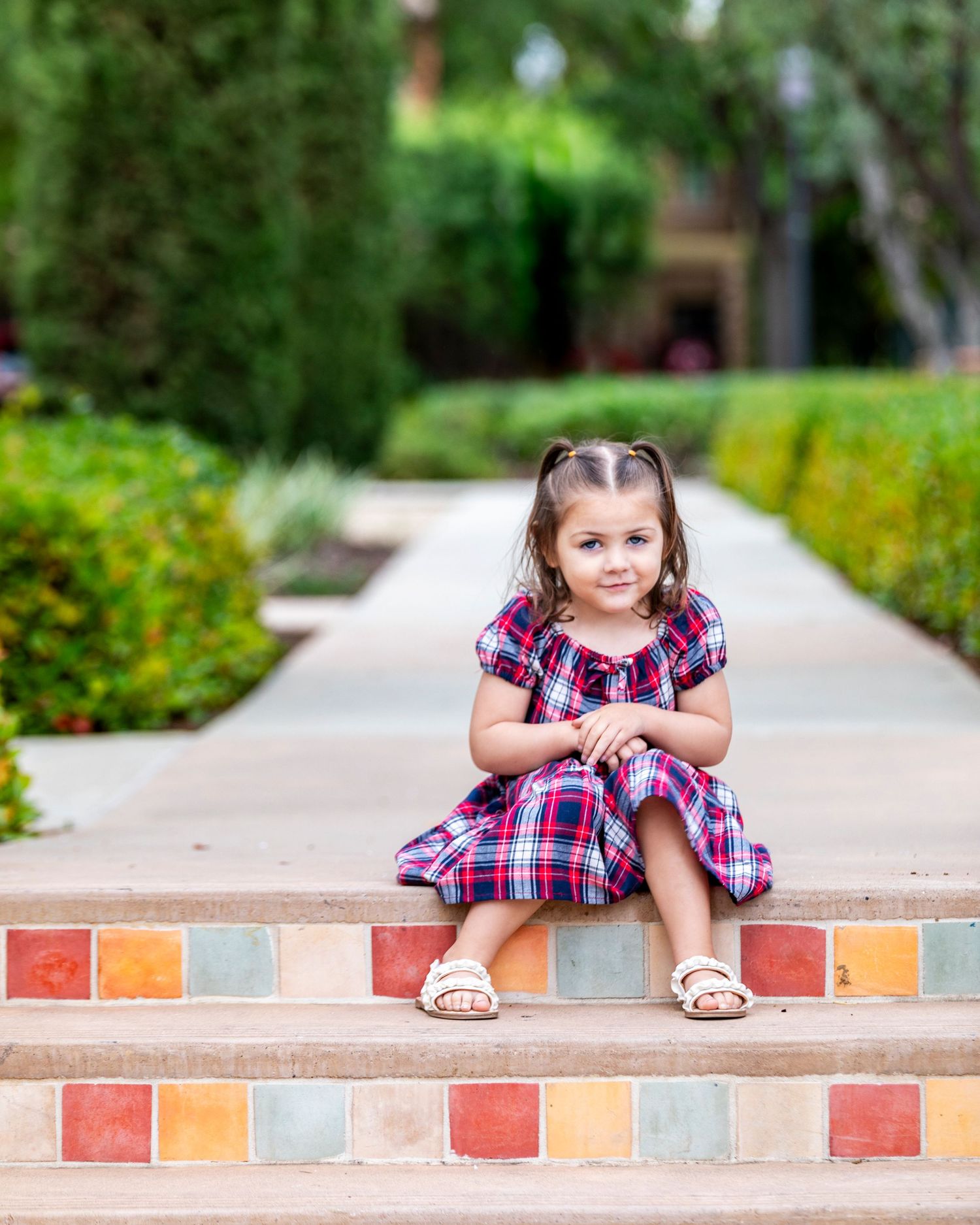 A young girl in a plaid dress sits on colorful tiled steps in a lush garden pathway.