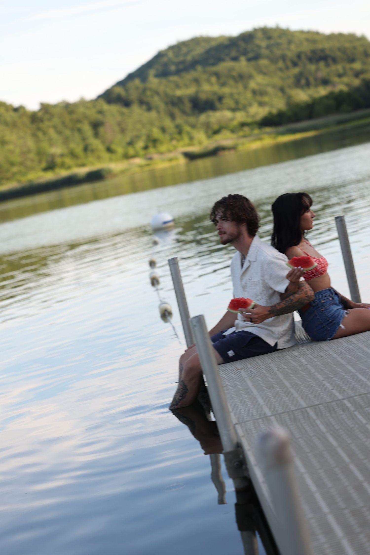 Couple sitting on a wooden dock eating watermelon by a calm lake with green hills in the background.