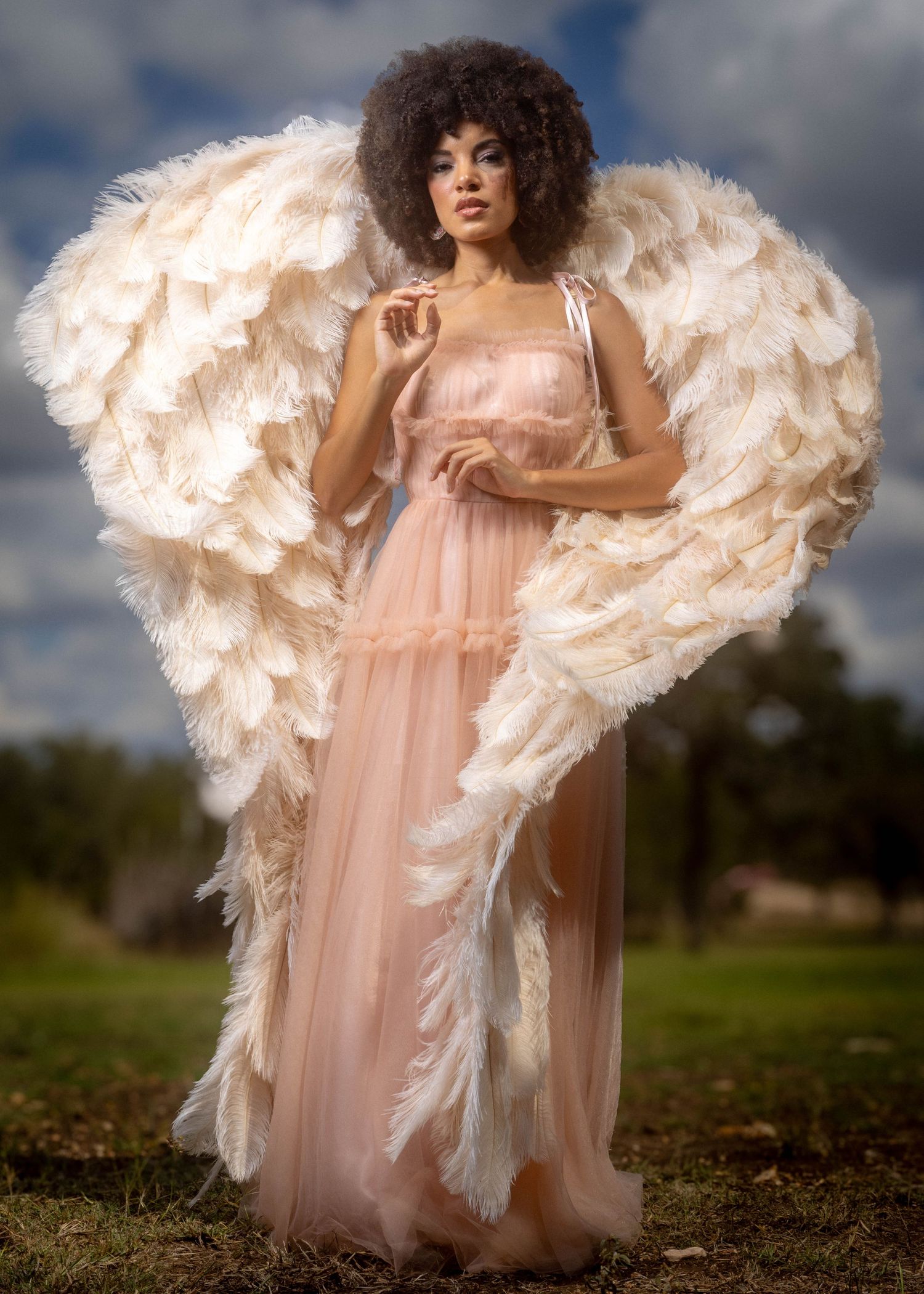 Woman in pink tulle gown with large white feather angel wings poses outdoors with curly afro hairstyle.