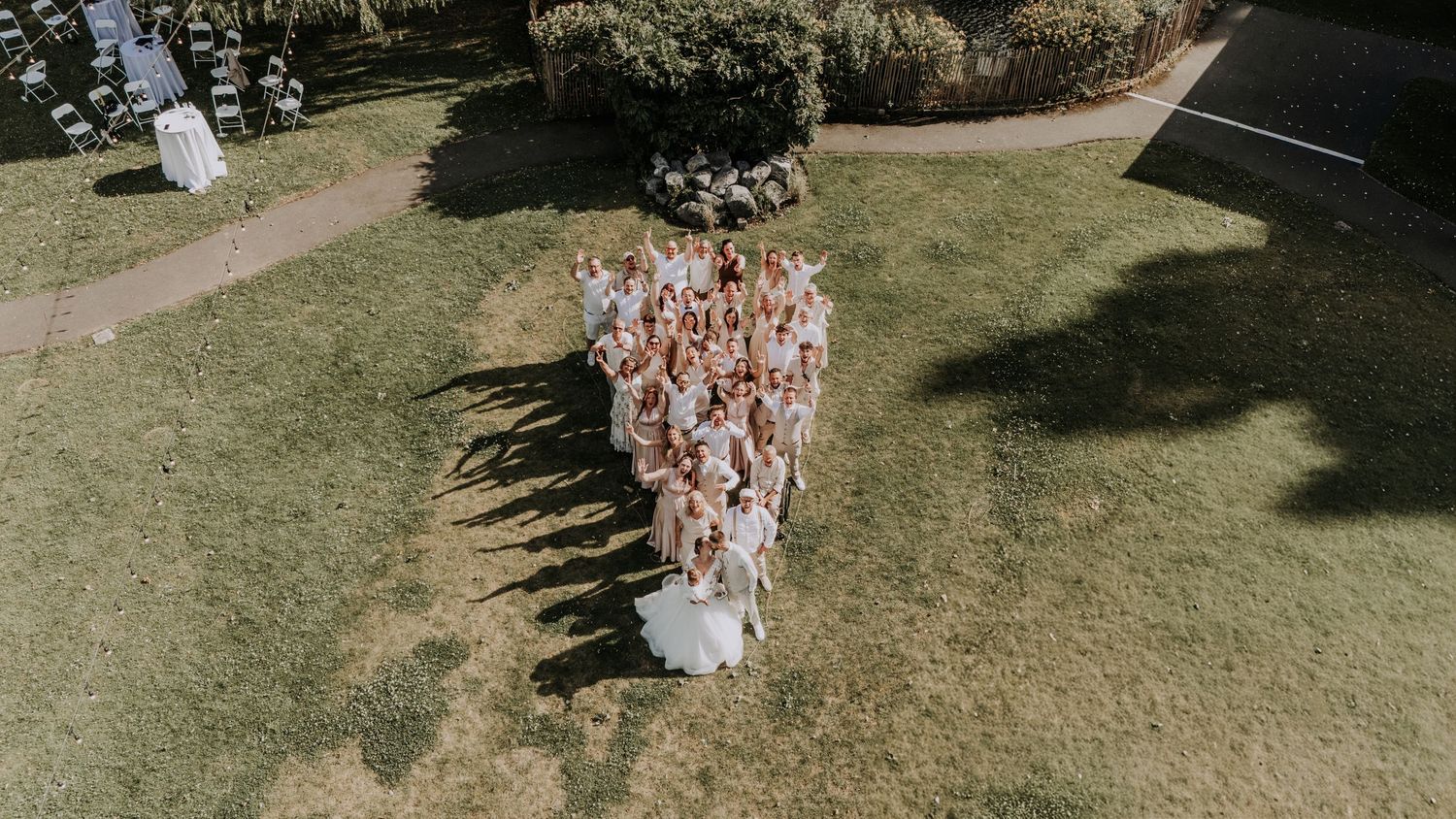 Aerial drone view of wedding party group photo on green lawn with long shadows cast on grass.
