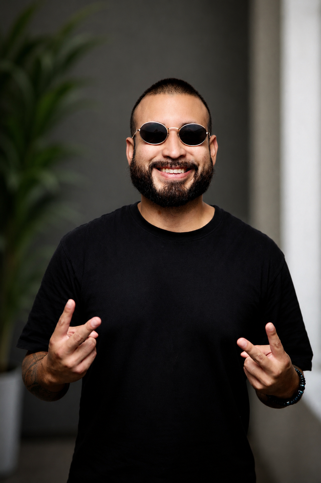 Smiling bearded man wearing black t-shirt and oval sunglasses making rock on hand gestures indoors.