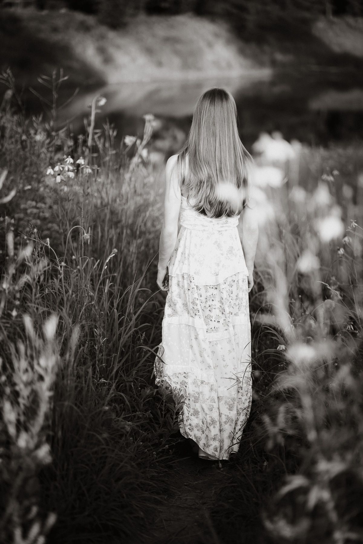 Black and white photo of a woman in a floral dress walking through a wildflower meadow toward a serene lake.