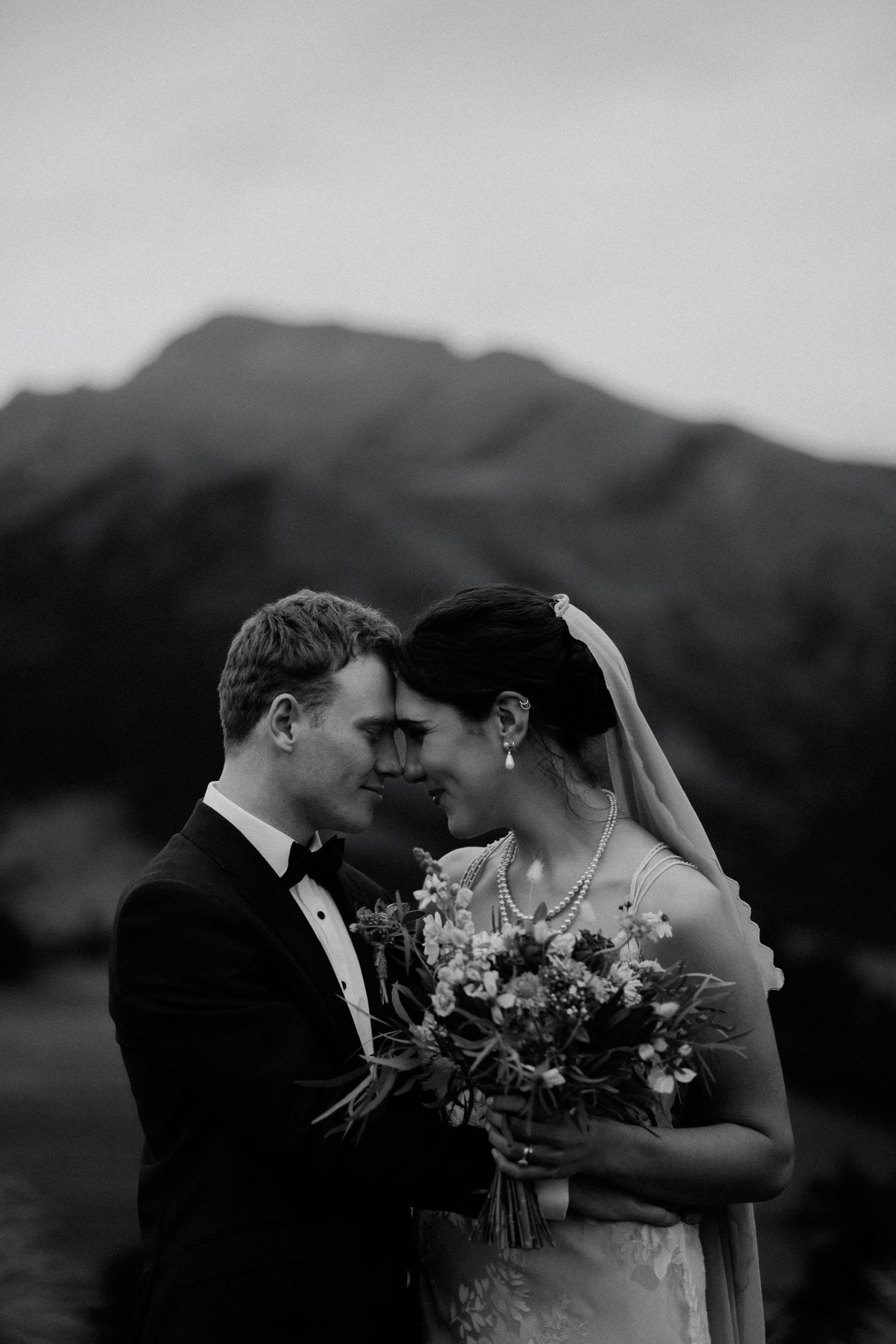 Black and white photo of a smiling bride in off-shoulder gown embracing groom in tuxedo, veil flowing in the wind.