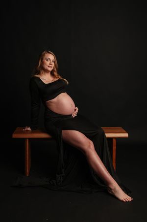 Pregnant woman in black off-shoulder gown sitting on wooden bench, cradling baby bump in professional maternity studio photo.