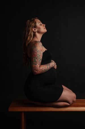 Pregnant woman with colorful tattoo sleeve sits gracefully on wooden bench in black outfit against dark background.