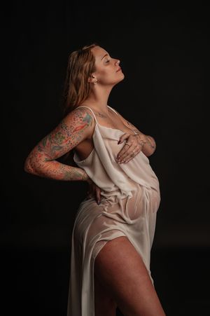 Pregnant woman in white silk dress with colorful tattoo sleeve posing against dark background.